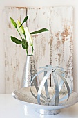 Sphere made of metal strips on metal dish and white lily in metal vase in front of vintage-style wooden board