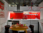 Plain white kitchen units with bright red splashback and red, stylised branches on wall unit doors, industrial ceiling and small dining area with set, wooden table in foreground