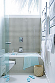 View through open glass door of bathroom with bathtub below window, beige wall tiles and stainless steel towel rail
