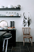 Tea towels with stag motif and old wooden chair next to crockery on wall-mounted shelves; designer bar stools at counter in foreground