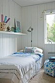 Bedspread on single bed below shelf on white, wood-clad wall in corner of room next to window