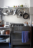 Modern gas cooker with stainless steel front next to table on castors and below pots and pans hanging from wall-mounted shelf