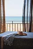 Strohhut auf Tagesbett vor offener Balkontür mit wehenden Vorhängen und Blick auf das Meer