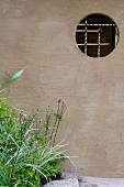 Wand im Garten mit Blick durch eine runde Öffnung auf geschnürte Bambusstäbe