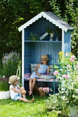 Blaues, offenes Gartenhäuschen mit Sitzbank und Regalfach in Sommergarten; sitzende Mädchen mit Strohhüten