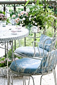 White metal chairs with seat cushions and bistro table in front of flowering plants in wire basket hanging on wrought iron balustrade