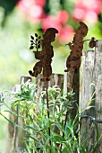 Patinated sheet metal garden gnomes on rods next to garden fence