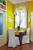 Bold yellow in corner of living room - tiny seating area with white covered chairs at wooden table below window