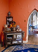 A wooden stove which has been converted to a china cupboard in front of an orange wall with a pointed arch passageway