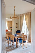 View through open door into dining room with Rococo chairs with blue seats in front of French windows with gathered curtains