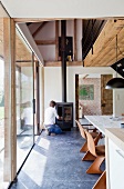 Wooden shell chairs at dining table in modern dining room with view of gallery; man stoking wood burner