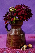 Dark red dahlias in classic ceramic jug against purple background; acorns on embroidered linen cloth
