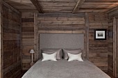 Schlafkammer mit Holzwänden und Holzdecke; in der Mitte ein Doppelbett mit gepolstertem Kopfende in schlichtem Grau