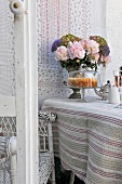 Cake under glass cover and vase of flowers on table covered in ethnic throw with wall hanging in background