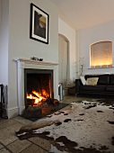 Large animal-skin rug on stone-flagged floor in front of burning open fire with leather sofa in background