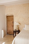 Antikes Nachtkästchen mit Tischlampe neben rustikaler Zimmertür in schlichtem Schlafzimmer