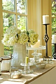 Porzellanvase mit weissen Hortensien und schmiedeeiserner Kerzenständer auf Tisch vor Fenster