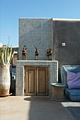Floor cushions and basketwork side table next to half-height, masonry cabinet with wooden doors on Mediterranean roof terrace