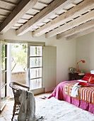 Schlafzimmer unter dem Dach - Rattanstuhl auf Flokatiteppich und Bett mit bunter Tagesdecke vor offener Balkontür