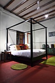 Four-poster bed with black frame on red floor in minimalist bedroom with wood beam ceiling