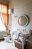 Waschtisch mit Keramikfüssen und Vintage Armatur an Wand mit rundem Spiegel, neben Fenster mit gerafftem, orangem Vorhang