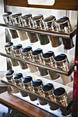 Dreireihiger, moderner Gewürzständer mit beschrifteten Metalldeckeln auf Glasflaschen