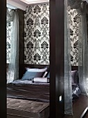 Schlafzimmer mit elegantem Doppelbett mit changierendem grau-violettem Überwurf und einer schwarz-weiss gemusterten Wandtapete