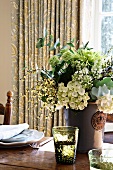 Bouquet in subdued colours in glazed ceramic vase and glasses with embossed pattern on rustic, set wooden table with floral curtain in background