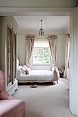 Feminine bedroom in soft shades of pink with old, white wardrobe, double bed with upholstered footer and pretty, gathered pale curtains in background. Elegant country-house ambiance.