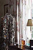 Wire tailor's dummy hung with innumerable necklaces next to antique dressing table with perfume bottles in front of floral curtains at window