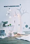 Stilisierter Baum mit Lichterkette neben Acrylglas Beistelltisch und Geschenketurm vor Wand mit Deko- Schneeflocken