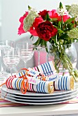 Rolled linen napkins with multicoloured stripes and name tags on stack of plates in front of summer bouquet in vase