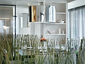 Looking over decorative plants to a hanging lamp over a dining table in front of white shelves