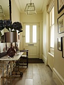 Table lamp and vase of silk flowers on vintage console table in narrow hallway of country house