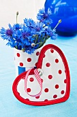 Heart-shaped pot holder in front of fabric-covered beaker of cornflowers