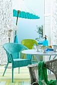 Set of wicker chairs in turquoise, green and white at round table; fan and parasol in front of sky blue wall in background