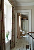 View from landing through open doorway into room with upholstered wicker armchair and antique, white wardrobe; floor-length curtains at windows add a pretty touch