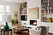 Modern painting over a fireplace, open bookshelves, marble top coffee table and white armchair in a living room