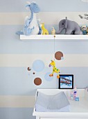 Nursery wall decorated with pale blue stripes and circles; various soft toys on floating shelf and pale blue changing mat on white changing unit