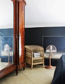 Large, antique mirrored cupboard with exotic wood veneer next to simple wicker chair and fan