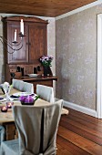 Antique bureau used as dining room dresser in room with subtle floral wallpaper; rustic dining table and chairs with loose linen covers in foreground