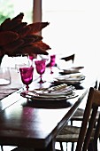 Magentafarbene Weingläser und exotischer Tischschmuck auf festlich gedecktem, antikem Holztisch mit einfachen Geflechtstühlen