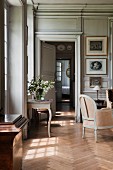 View through open interior doors of grand salon with antique side table below window and delicate armchair on herringbone parquet floor