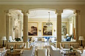Spacious hotel lobby with classic, pale upholstered furniture and landscape paintings separated by Ionic stone pillars