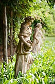 Statue of girl in flowing gown with basket of flowers in row of antique statuary amongst luxuriant green of romantic hotel gardens