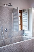 Badewanne und Wand mit hellen Mosaikfliesen und offene Innenläden am Fenster in modernem Bad
