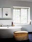 Polsterhocker mit goldgelbem Bezug und Fransen vor moderner Badewanne neben Sprossenfenster