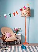 Bunting, standard lamp with orange-painted base and modern floral lampshade casually combined with vintage armchair against pale blue wall