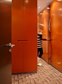 Abstellraum mit orangen raumhohen Einbauschränken und grau gefliestem Boden, eine schwarz-weiß gekleidete Frau öffnet einen Schrank