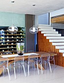 Acrylglas-Stühle an massivem Holztisch; im Hintergrund ein Weinregal und eine offene Treppe ohne Geländer